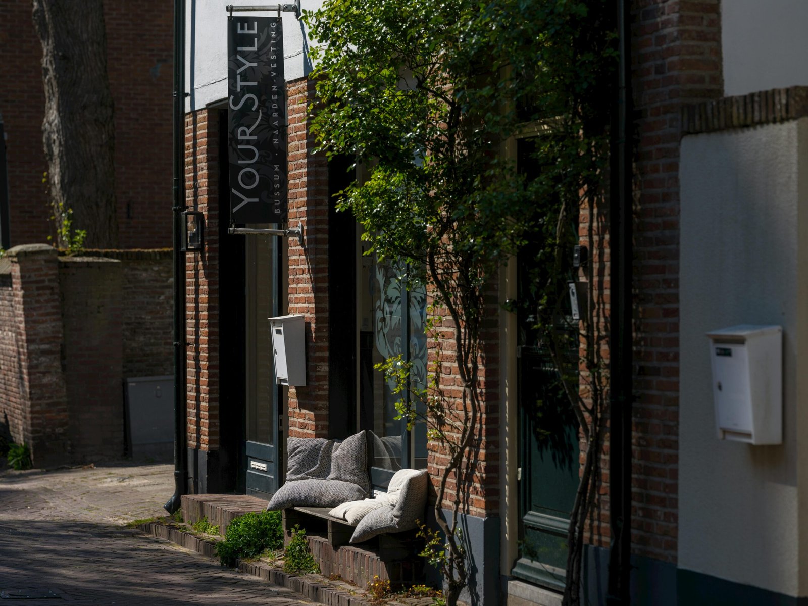 Outdoor seating with pillows outside a stylish brick-walled salon in an urban setting, enveloped by greenery.